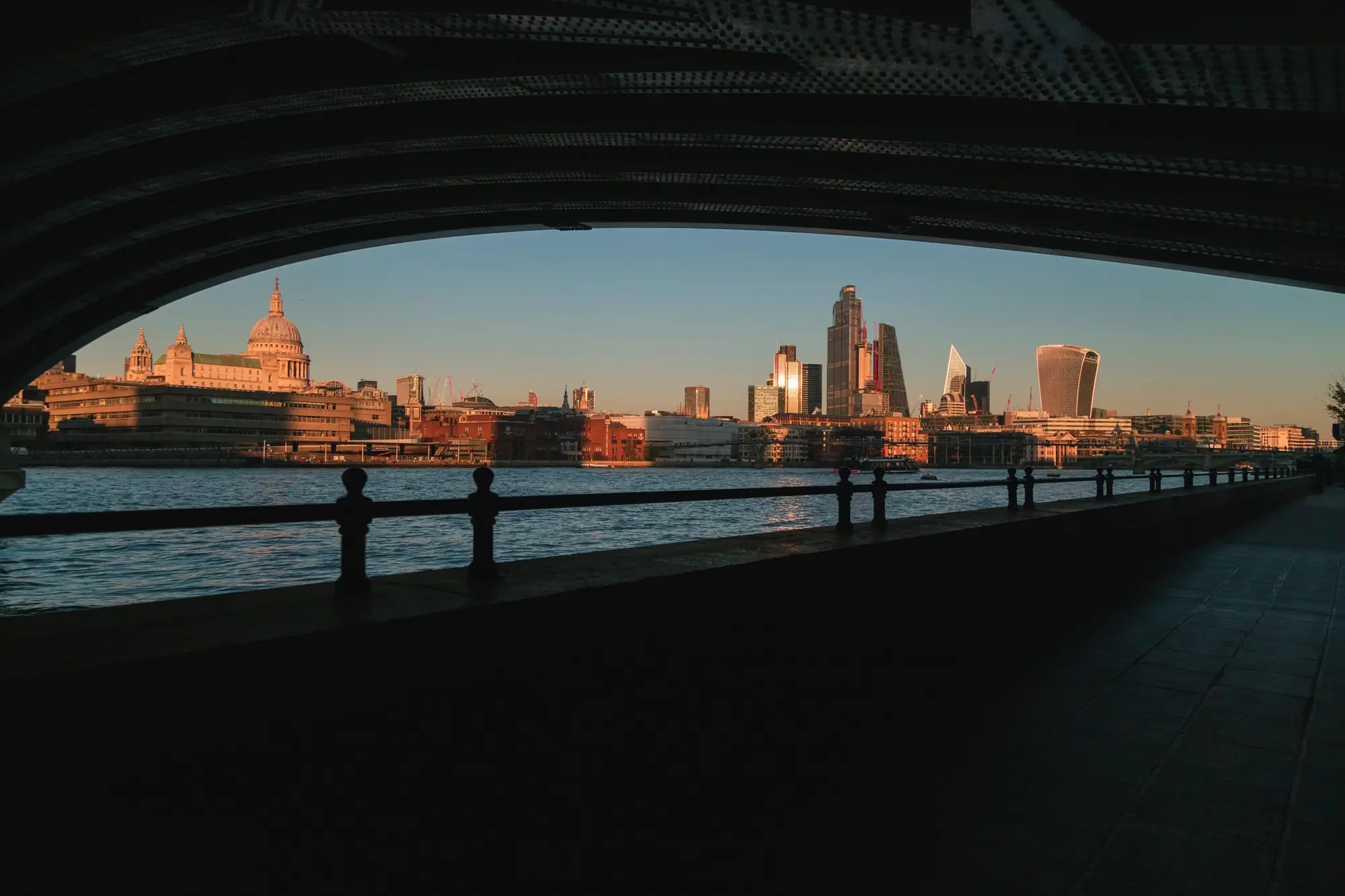 The City of London skyline at dusk, seen from beneath a bridge arch across the Thames.