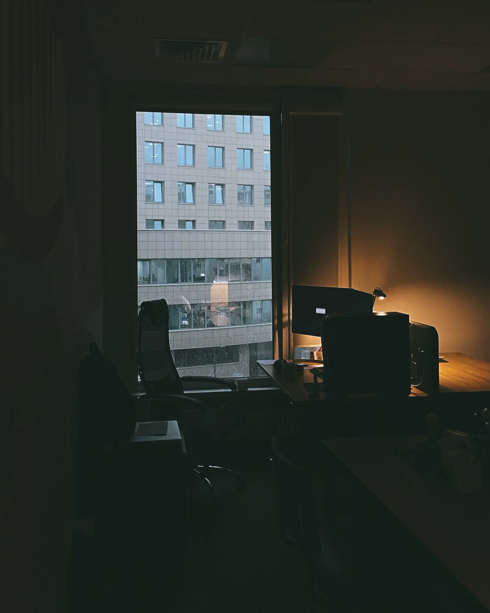 A dimly lit office at a desk by a window, one monitor awake