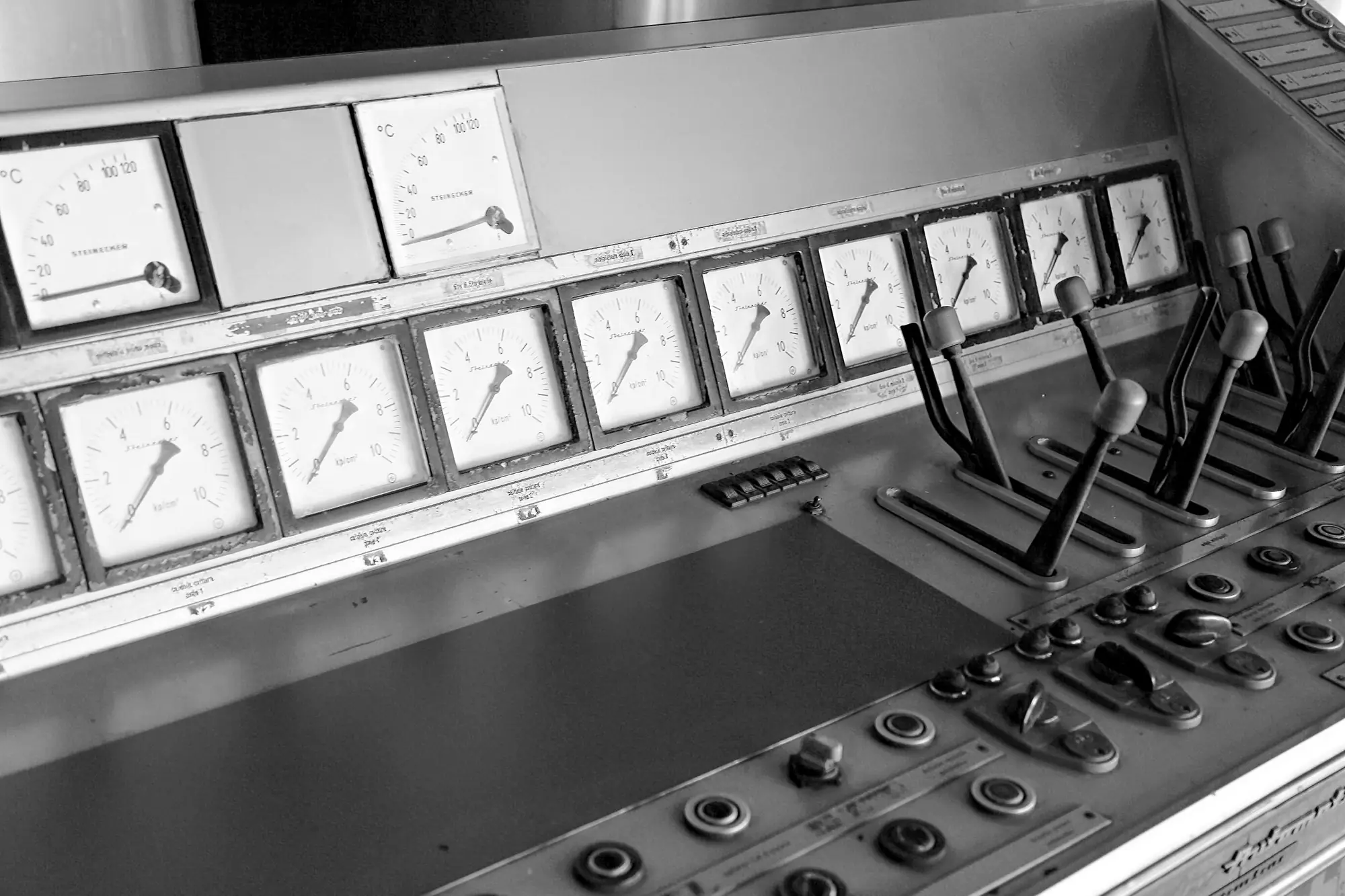 A bank of analogue gauges and levers across an industrial control desk, in black and white.